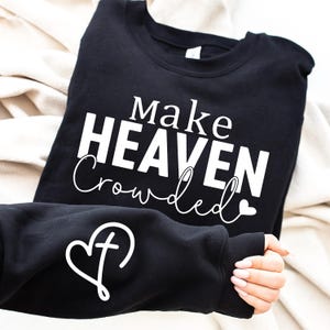 May include: Black sweatshirt with the text "Make HEAVEN Crowded" in white, with a heart and cross design on the sleeve. The sweatshirt is folded on a cream-colored surface.