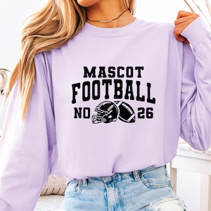 May include: A light purple long-sleeve sweatshirt with the words "MASCOT FOOTBALL NO. 26" in black block letters. A black football helmet and football are printed below the text. The sweatshirt is made of a soft material.