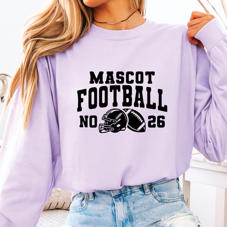 May include: A light purple long-sleeve sweatshirt with the words "MASCOT FOOTBALL NO. 26" in black block letters. A black football helmet and football are printed below the text. The sweatshirt is made of a soft material.