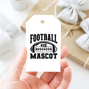 May include: A white gift tag with a black football graphic and the words "FOOTBALL MASCOT #15". The tag is held in a hand, with a blurred background of gift wrapping and bows.