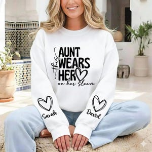 May include: White sweatshirt with the text "Aunt Wears Her Heart on Her Sleeve." The sleeves feature heart designs with the names "Sarah" and "David." The sweatshirt is a casual, comfortable style.