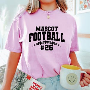 May include: A light pink t-shirt with the words "MASCOT FOOTBALL #26" in black. The design includes a football graphic. A yellow and white mug with a smiley face is held in the hand.