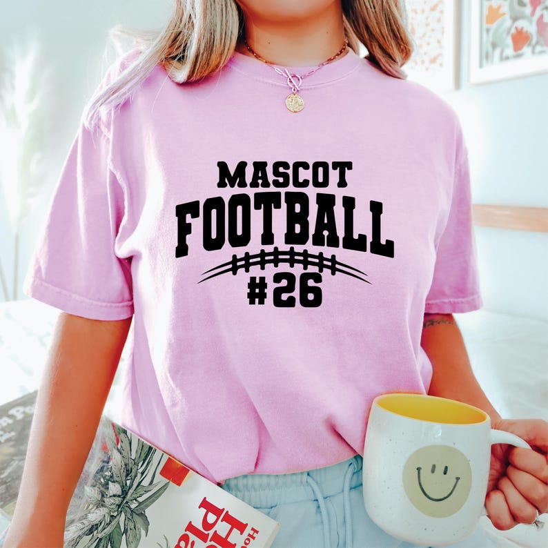May include: A light pink t-shirt with the words "MASCOT FOOTBALL #26" in black. The design includes a football graphic. A yellow and white mug with a smiley face is held in the hand.