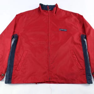 May include: A red zip-up jacket with navy blue accents on the sleeves and sides. The jacket has a small rectangular patch on the chest and two front pockets. The collar and cuffs are navy blue.