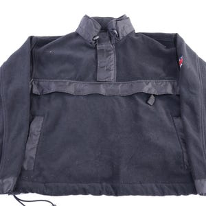 May include: A dark navy blue fleece pullover jacket with a high collar and a small pocket on the front. The jacket has black accents and a small emblem on the upper left sleeve. The bottom hem has a drawstring.