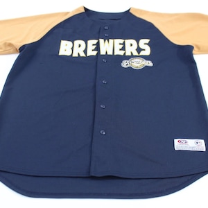 以下が含まれることがあります： ネイビーブルーの野球ジャージ。袖はタン色。胸には白いブロック体で「BREWERS」の文字。ロゴが「BREWERS」の上にあります。ボタン留めのジャージです。