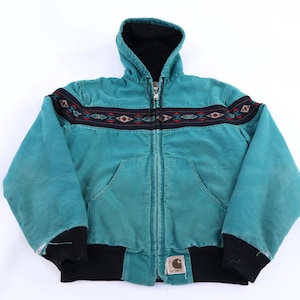 May include: Teal hooded jacket with a black hood lining and cuffs. The jacket features a zip-up front and two front pockets. A decorative band with geometric patterns in red, black, and white accents the chest area. A Carhartt label is visible.