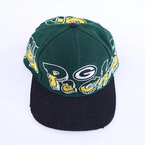 Peut inclure: Une casquette de baseball vert foncé avec une visière plate noire. La calotte présente un grand texte stylisé blanc et jaune indiquant "PACKERS" et inclut un logo "G" blanc et vert. La casquette a une structure à six panneaux avec un bouton.