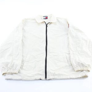 May include: Off-white jacket with a black zipper, two front pockets, and a collar. The jacket has long sleeves with elastic cuffs and a small logo on the chest. The jacket is made of a lightweight material.