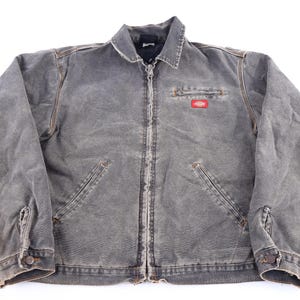 May include: A charcoal gray zip-up work jacket with a distressed finish, featuring a fold-down collar, long sleeves with buttoned cuffs, two slanted front pockets, and a small chest pocket with a red and yellow Dickies brand logo patch.
