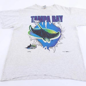 May include: A light gray t-shirt featuring the words "TAMPA BAY" in purple block letters above a graphic of a manta ray. The manta ray is black and green, set against a blue and green background.