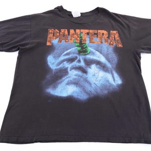 May include: A black short-sleeve t-shirt featuring the word PANTERA in orange block letters at the top. Below the text is a graphic of a blue, hazy, skull-like shape with a green, clawed hand reaching down toward the center of the forehead.