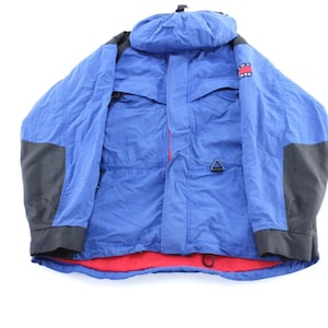 May include: A blue hooded jacket with black accents on the shoulders and sleeves. The jacket has a red interior and a small logo patch on the upper chest. It features multiple pockets and a drawstring at the bottom.