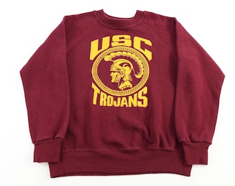 May include: Maroon crewneck sweatshirt with yellow USC Trojans logo. The logo features a Trojan warrior head in a circular design with the text "USC" above and "Trojans" below. The sweatshirt has ribbed cuffs and hem.