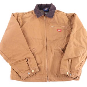 May include: A tan canvas work jacket featuring a dark brown corduroy collar, a full-length front zipper, and long sleeves with buttoned cuffs. The jacket includes a zippered chest pocket with a red Dickies brand label and two lower hand pockets.