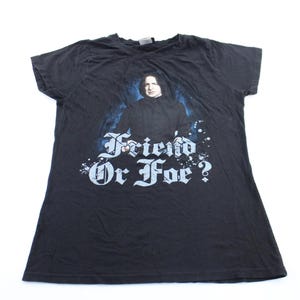 May include: A black short-sleeved t-shirt featuring a graphic print of a person with dark hair wearing a black robe. Below the figure, the text "Friend Or Foe?" is printed in a white, stylized gothic font against a blue and black background.