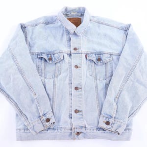 May include: A light-wash blue denim jacket featuring a classic collar, long sleeves with buttoned cuffs, and a front button closure. The garment includes two chest pockets with buttoned flaps and a brown leather-like brand patch at the inner neck.