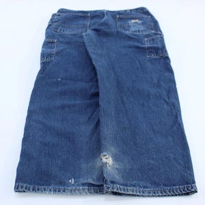 Puede incluir: Pantalones de trabajo de mezclilla azul con múltiples bolsillos y un área desgastada cerca de la rodilla. Los pantalones están colocados planos sobre una superficie blanca, mostrando los bolsillos traseros y el diseño general.