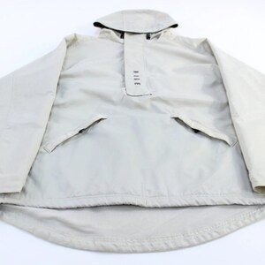 May include: A beige hooded pullover jacket featuring long sleeves, a front placket with the text NIKE printed vertically, and two zippered pockets. The hem includes an adjustable drawstring with black toggles for a secure fit at the waist.