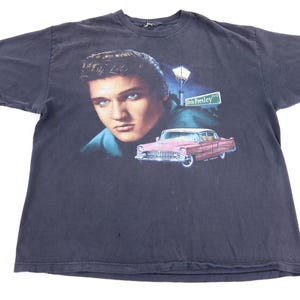 May include: A black short-sleeve t-shirt featuring a graphic print of Elvis Presley's face, a pink vintage car, a street lamp, and a green street sign that reads Elvis Presley Blvd. The shirt has a crew neckline and a standard fit.