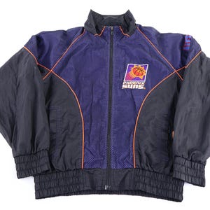 May include: A vintage Phoenix Suns basketball team jacket. The jacket is primarily black with a purple center panel featuring an orange and purple team logo. Orange trim outlines the purple panel. The jacket has a full zipper and ribbed cuffs and hem.