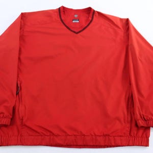 May include: A bright red pullover windbreaker with a V-neck collar and long sleeves. The windbreaker has a black trim around the collar and cuffs. The bottom hem is elasticized. The jacket is made of a lightweight material.