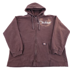 May include: A brown zip-up hoodie featuring a hood with matching drawstrings, long sleeves, and two front pockets. The chest displays the white text Dickies with a brown wing graphic. A small Dickies brand label is attached to the front pocket.