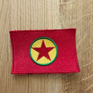 May include: A rectangular red patch featuring a central design. The design includes a yellow circle with a red star in the center, outlined in green. The patch is likely an emblem or insignia.
