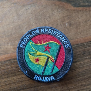 May include: A round black and white patch with the text "People's Resistance" in a circle around a green, yellow, and red flag with a red star. The text "Rojava" is below the flag.
