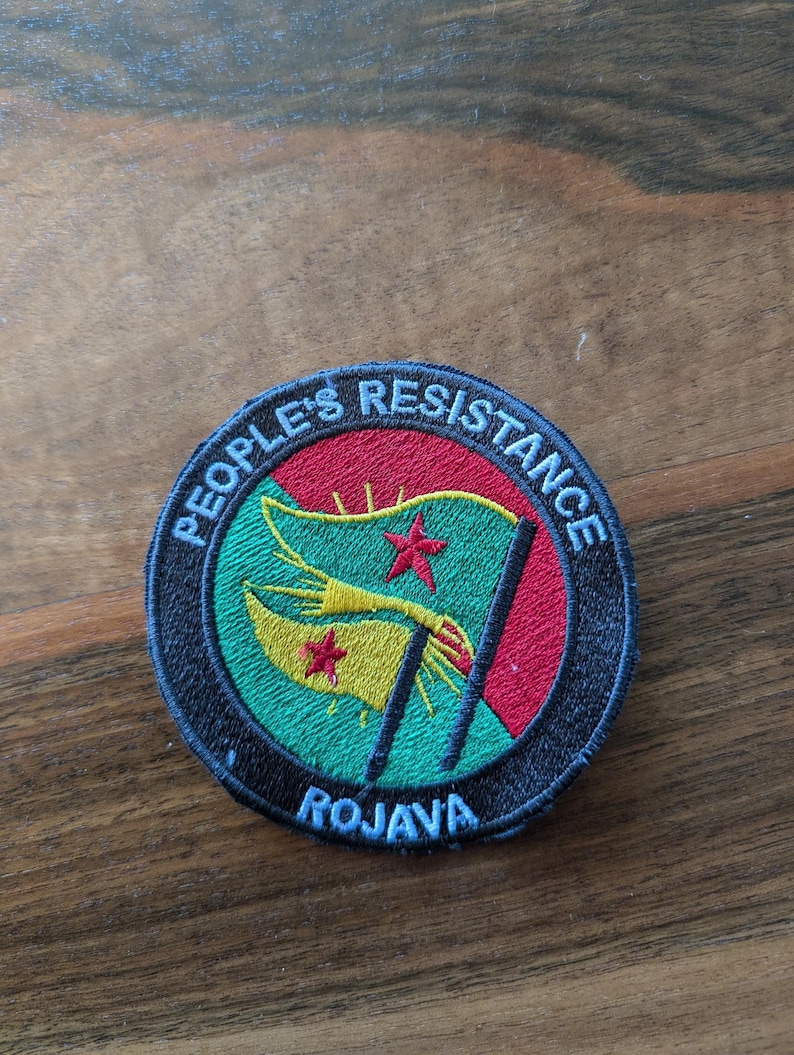 K&ouml;nnte beinhalten: Ein runder schwarz-wei&szlig;er Aufn&auml;her mit dem Text "People's Resistance" in einem Kreis um eine gr&uuml;n-gelb-rote Flagge mit einem roten Stern. Der Text "Rojava" befindet sich unter der Flagge.