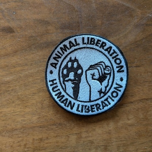 May include: A white and black embroidered patch with the text "Animal Liberation - Human Liberation" and a raised fist and paw print.