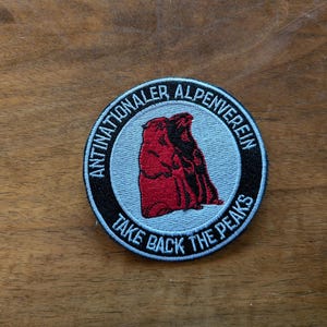 Könnte beinhalten: Ein schwarzer und weißer runder Patch mit dem Text "Nationaler Alpenverein" und "Take Back the Peaks" in Weiß. Der Patch zeigt eine rote und weiße Illustration von zwei Murmeltieren.