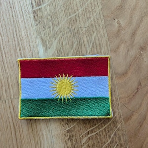 May include: Embroidered patch featuring the flag of Kurdistan. The rectangular patch has horizontal stripes of red, white, and green, with a central gold sun emblem. The patch is outlined with gold stitching.
