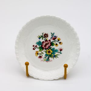 May include: A white ceramic plate with a scalloped edge and a floral design. The design features a bouquet of flowers in shades of red, yellow, blue, and green. The plate is displayed on a gold-coloured stand.