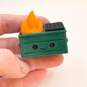 May include: A green plastic toy dumpster with a smiling face and black eyes. The dumpster has a black lid and a bright orange flame on top.