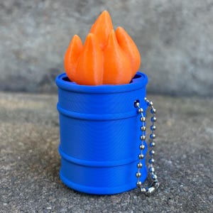 May include: A blue cylindrical barrel-shaped container with a bright orange flame-shaped insert protruding from the top. A silver-toned metal ball chain is attached to the side of the blue barrel, allowing it to function as a keychain or hanging charm.