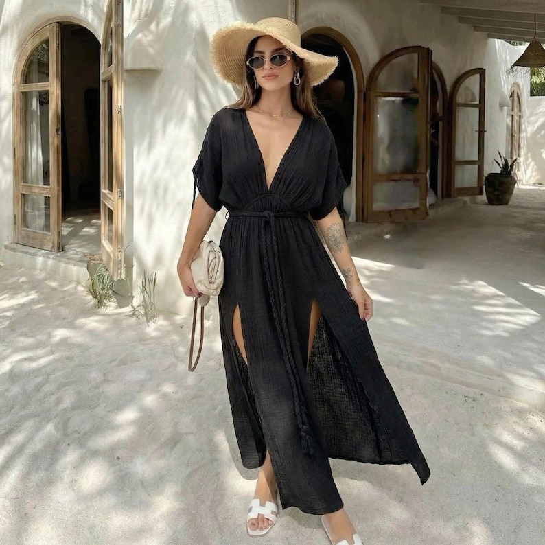 Puede incluir: Un vestido largo negro con escote pronunciado y aberturas laterales altas. El vestido est&aacute; ce&ntilde;ido a la cintura con un cintur&oacute;n a juego. La persona lleva un sombrero de paja, gafas de sol y sandalias blancas. Se sostiene un peque&ntilde;o bolso blanco en la mano.