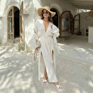 Puede incluir: Una mujer con un vestido largo blanco de lino con escote pronunciado y aberturas laterales altas. Lleva un sombrero de paja, gafas de sol y sandalias blancas. Sostiene un peque&ntilde;o bolso estampado. El vestido tiene un cintur&oacute;n a juego.