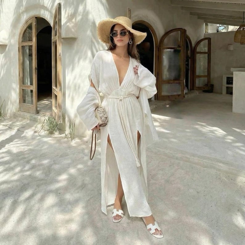Puede incluir: Una mujer con un vestido largo blanco de lino con escote pronunciado y aberturas laterales altas. Lleva un sombrero de paja, gafas de sol y sandalias blancas. Sostiene un peque&ntilde;o bolso estampado. El vestido tiene un cintur&oacute;n a juego.