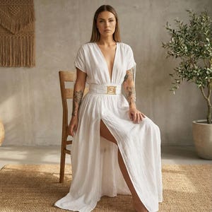 May include: A white linen maxi dress with a plunging neckline and a high slit. The dress features short sleeves and a wide belt with a gold-colored square buckle. The model is seated on a wooden chair.