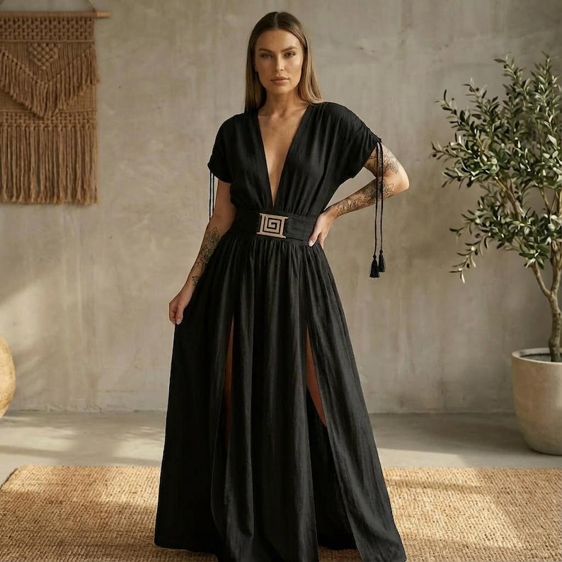 May include: A black maxi dress with a plunging neckline and short sleeves. The dress features a gold-colored belt with a geometric buckle and high slits on both sides. Tassels hang from the sleeves. The dress is made of a flowing fabric.