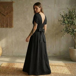 May include: A black maxi dress with a deep V-neckline and short sleeves. The dress features a fitted bodice with a smocked waist and a flowing skirt. A bow detail is at the back. The dress is made of a lightweight fabric.