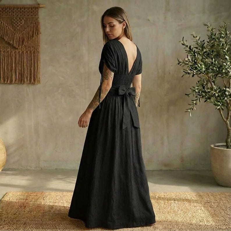 May include: A black maxi dress with a deep V-neckline and short sleeves. The dress features a fitted bodice with a smocked waist and a flowing skirt. A bow detail is at the back. The dress is made of a lightweight fabric.