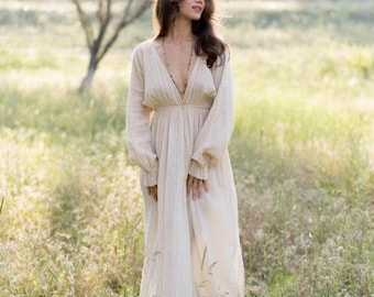 Puede incluir: Un vestido largo color crema con cuello en V profundo y mangas largas. El vestido tiene una silueta fluida y está hecho de una tela ligera. Las mangas tienen un ligero volante en la muñeca. El vestido se usa al aire libre en un campo de hierba alta.