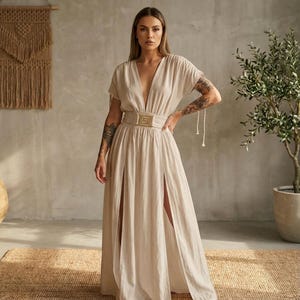 May include: A beige maxi dress with a plunging neckline and short sleeves. The dress features a high waist with a decorative belt and two high slits in the front. The dress is made of a lightweight fabric and has a flowing silhouette.