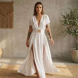 May include: A white maxi dress with a plunging neckline and short sleeves. The dress features a high waist with a gold-colored buckle belt and high slits on both sides. The dress is made of a lightweight fabric.