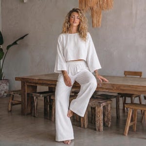 May include: A two-piece set consisting of a white long-sleeved cropped top with a round neckline and matching wide-leg trousers. The garment is made of a lightweight, textured fabric that falls loosely, creating a relaxed and comfortable silhouette.