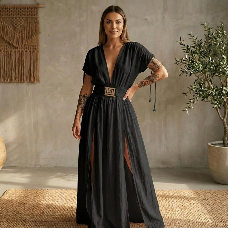 May include: A black maxi dress with a plunging neckline and short sleeves. The dress features a gold-colored buckle belt and high side slits. The model is standing in front of a neutral-toned wall and a woven rug.