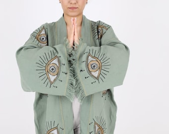 May include: A sage green kimono with a fringed hem and embroidered eye designs. The kimono is open in the front and the sleeves are long.
