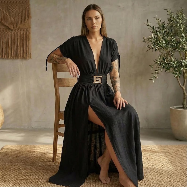 May include: A black maxi dress with a deep V-neck and a high slit. The dress features short sleeves with tie details and a gold-colored belt with a geometric buckle. The model is seated on a wooden chair.